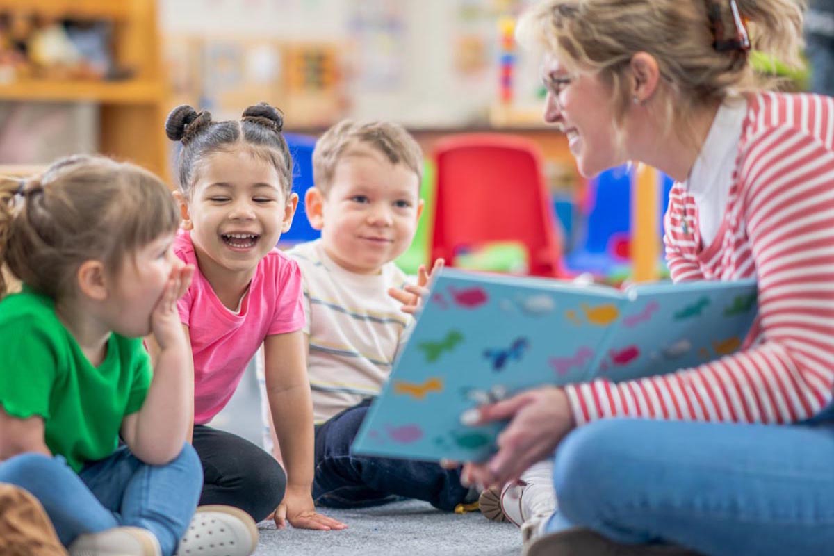A preschool teacher sits on the floor of her classroom with a small group of students as she reads them a book. 