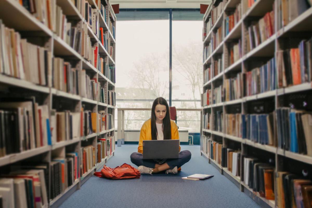 Teenage girl using laptop while sitting beside bookshelves in library